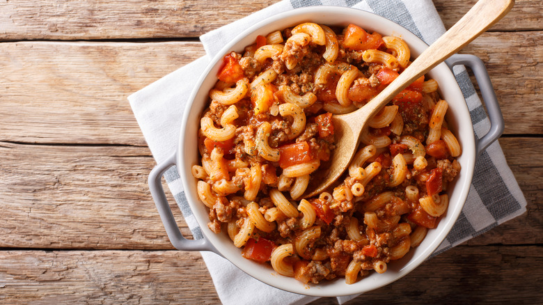 A pot of American Goulash on a wooden surface with a wooden spoon stuck inside