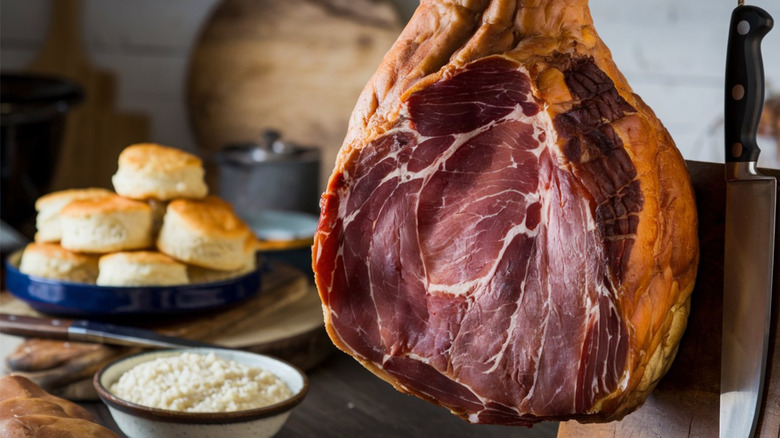 A country ham hanging in the kitchen with biscuits and grits in the background