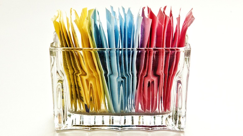 A glass container with several types of sweetener packets inside