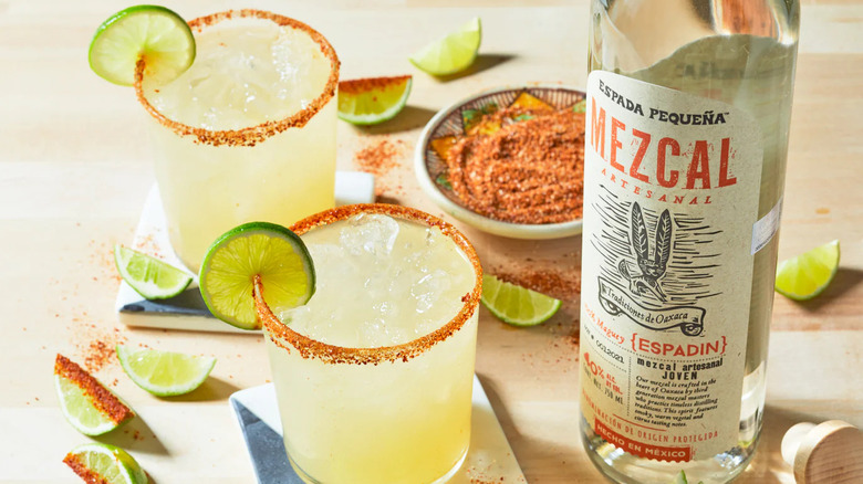 A bottle of Espada Pequeña mezcal next to two margaritas on the rocks with lime garnishes and salted rims
