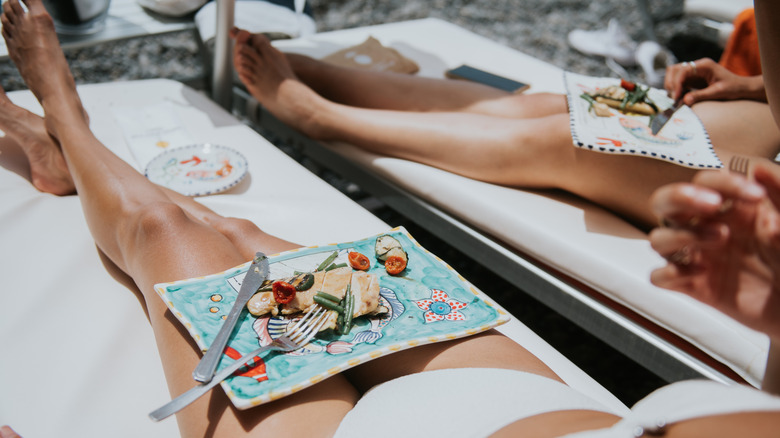 Two women lounging on sunchairs in swimsuits, with plates of food
