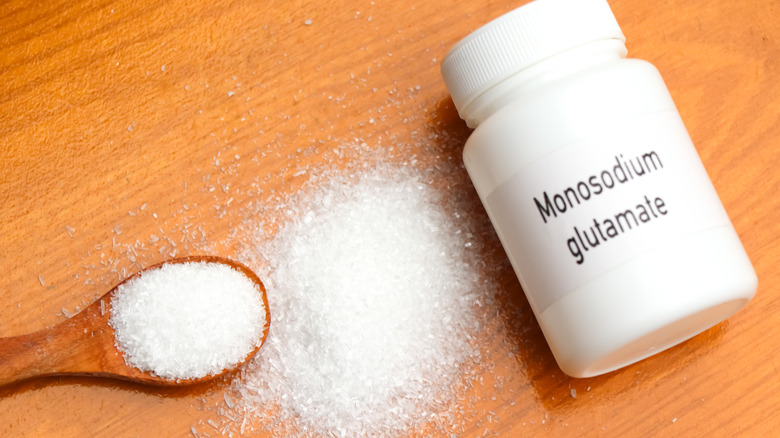 A bottle of MSG on a wooden table with a heap and a spoonful of MSG on the side