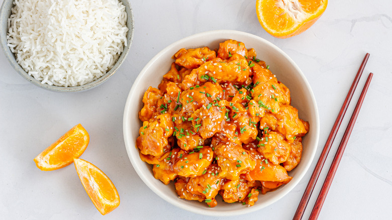 Orange chicken in a bowl with a bowl of rice, chopsticks, and orange slices on the side on a white table