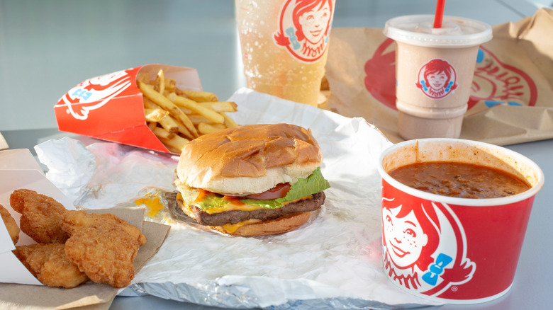 An assortment of Wendy's items, including a Dave's single cheeseburger, chicken nuggets, chili, fries, soda, and a Frosty against a gray background.