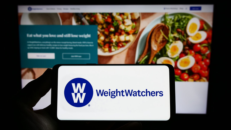 A phone showing the Weight Watchers logo with a computer screen in the background.
