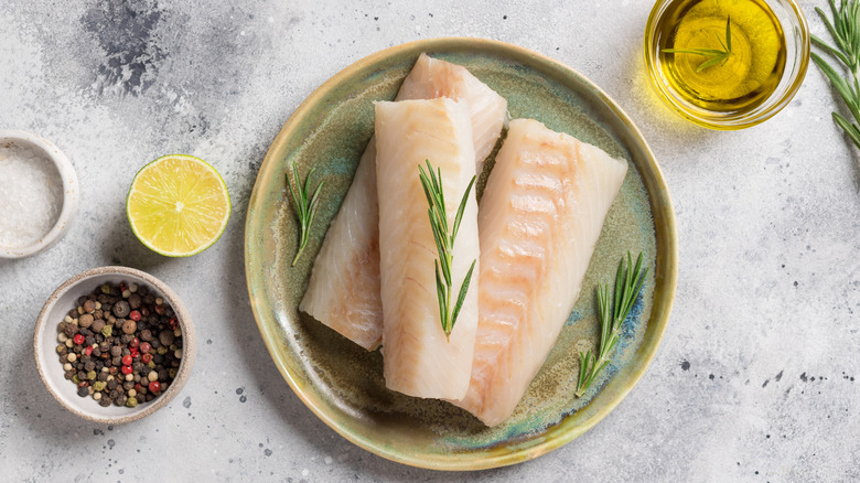 A plate with three cod filets surrounded by lemon, oil, herbs, and pepper
