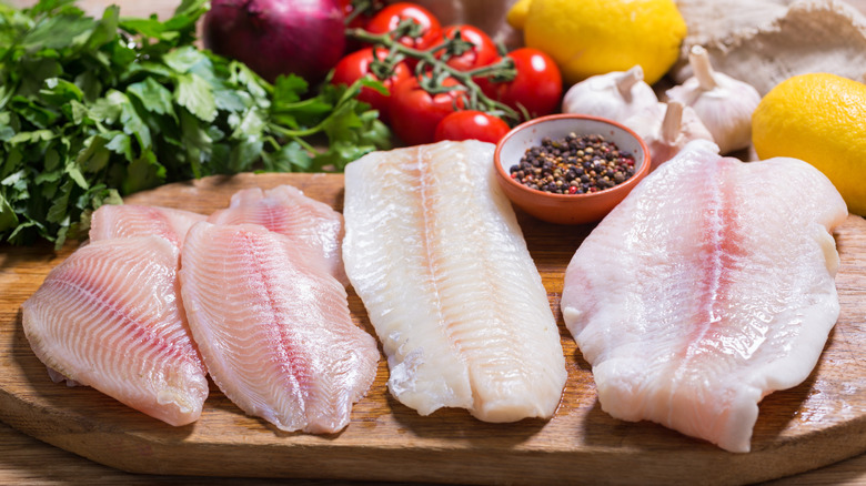 Four raw catfish filets served on a wood plank and surrounded by spices, herbs, and vegetables