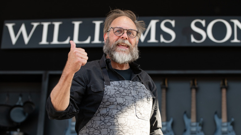 a bearded Alton Brown gives a thumbs up while wearing a gray apron with various foods and cookware items on it