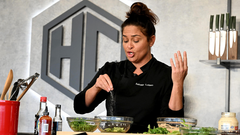 Chef Antonia Lofaso stands behind a counter, salting a bowl of green veggies while talking to an audience