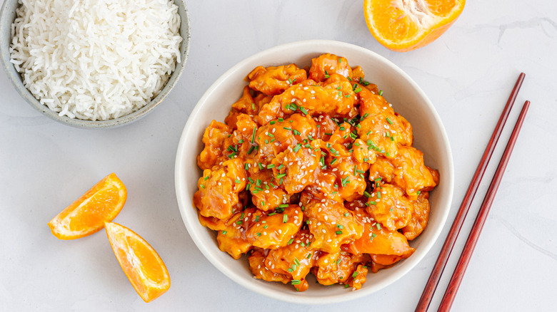 A bowl of orange chicken sits next to a bowl of rice, with orange segments and chopsticks