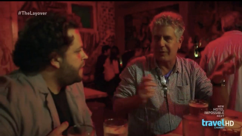 Anthony Bourdain and a friend talk with their hands in a dimly lit bar.