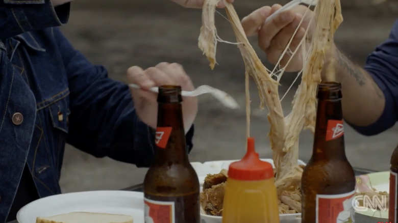 Two men using forks to separate an especially long piece of pulled pork, with two beers in the foreground.