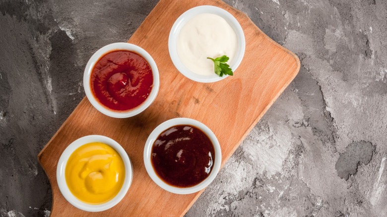 An array of condiments cups on a wooden cutting board