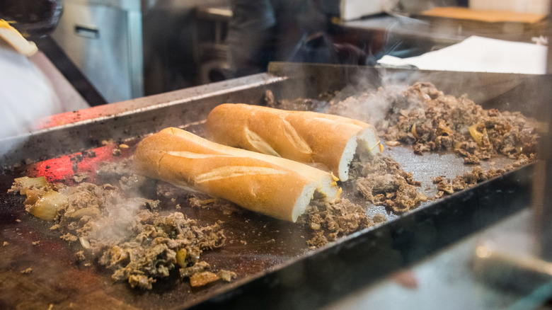 A flat-top grill full of shaved steak for cheesesteak, with two rolls ready to be flipped for sandwiches