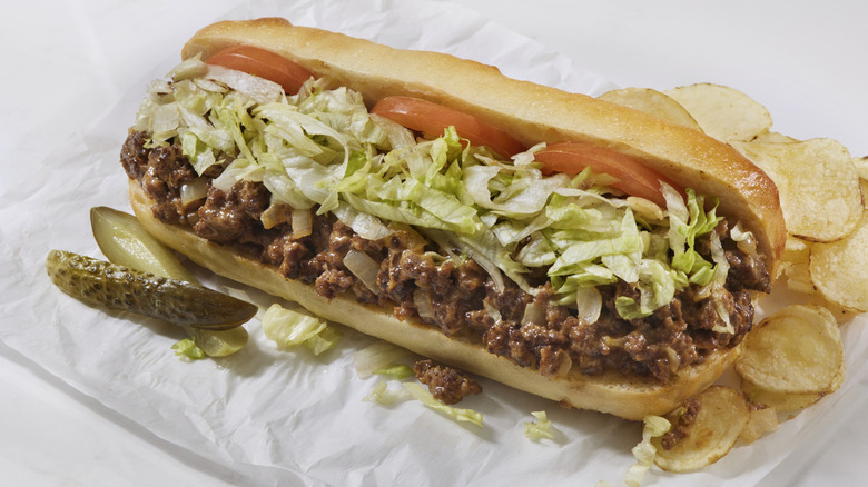A cheesesteak with lettuce and tomato, and a pickle and potato chips on the side