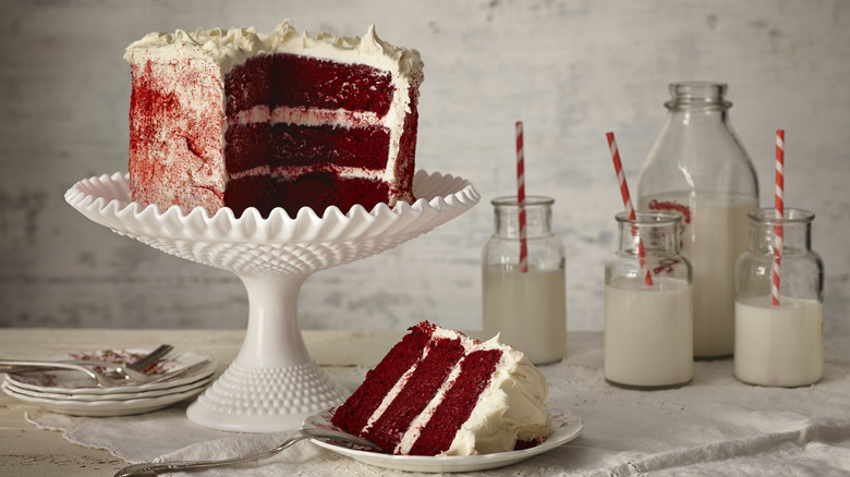 A red velvet cake on a stand with glasses of milk in the background
