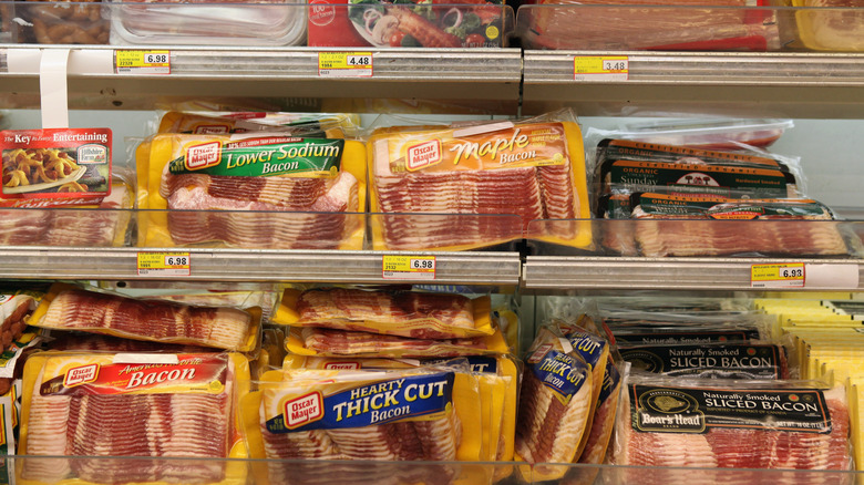 rows of different kinds of bacon on grocery store shelves.