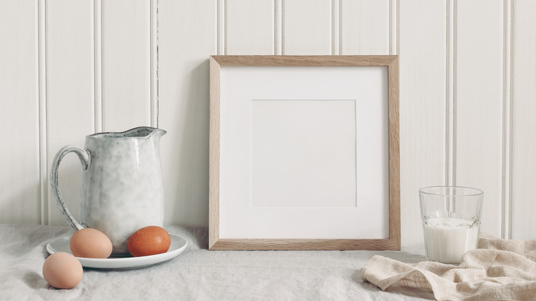 A kitchen still life of empty photo frame, eggs and milk in front of beadboard wall