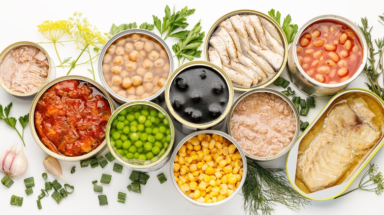 Several opened food cans, containing things like black olives, green peas, tuna, and anchovies, all surrounded by fresh herbs.