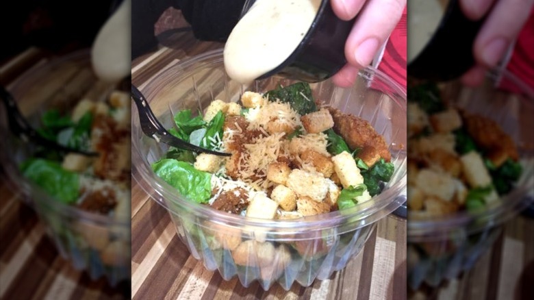 Plastic takeout bowl of Chicken Caesar Salad with a hand pouring dressing over it