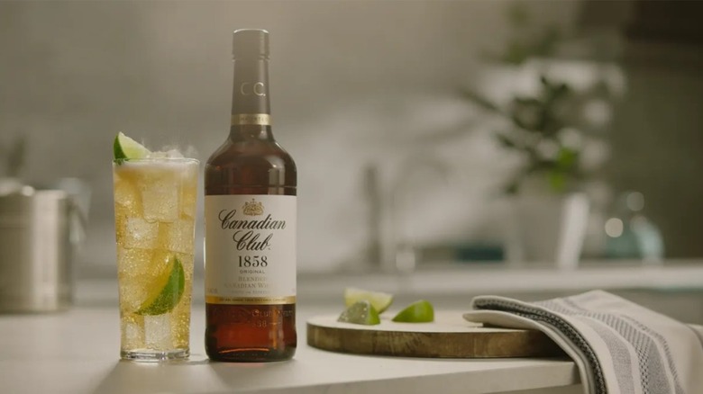 A bottle of Candian Club 1858 next to a cocktail and a cutting board with limes
