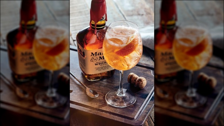 A bottle of Maker's Mark whiskey next to a cocktail on a wooden cutting board