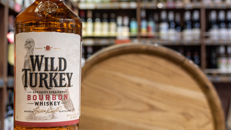 A bottle of Wild Turkey bourbon next to a whiskey barrel with rows of bottles in the background