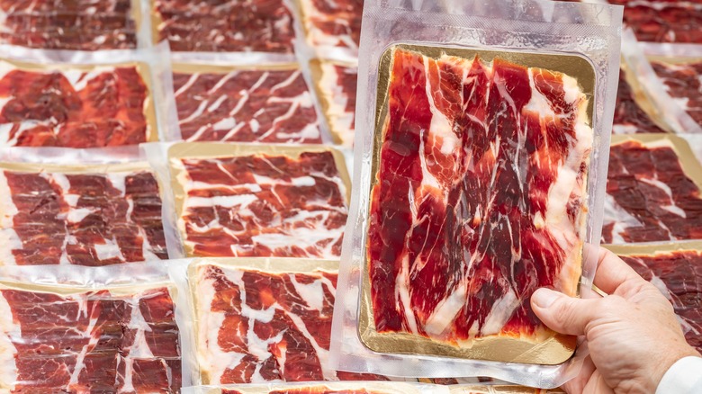 A person holds a package of sliced ham in front of a larger display of the meat