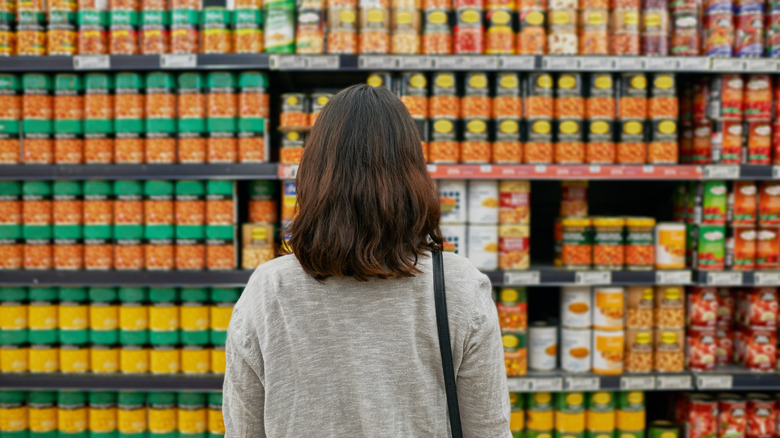 the back of a woman's head and torso as she gazes at canned foods at the grocery store