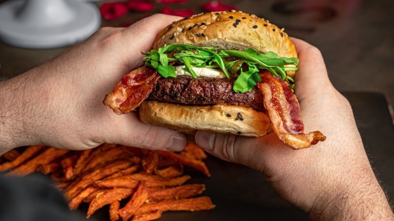 The Works bacon burger in two hands with fries in the background