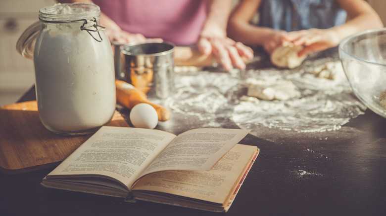 A vintage cookbook on a kitchen table, surrounded by baking ingredients, as a girl and her grandmother make dough in the background.