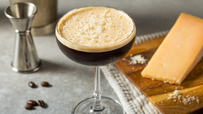 An espresso martini dusted with grated Parmesan, with a Parmesan wedge and jigger in the background