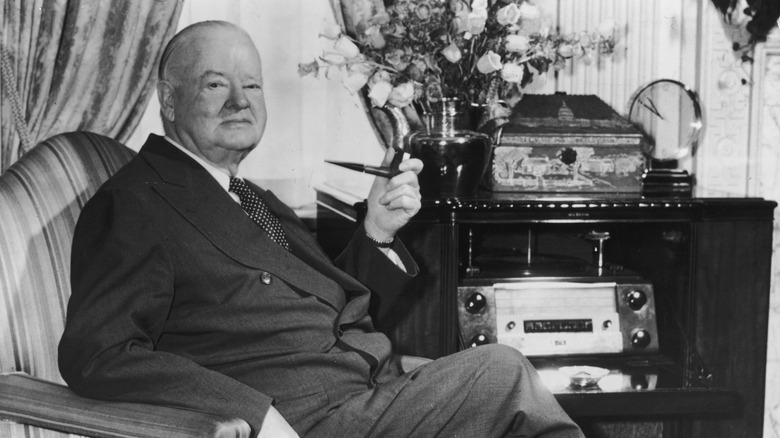 Black-and-white photo of President Herbert Hoover sitting in a chair and smoking a pipe