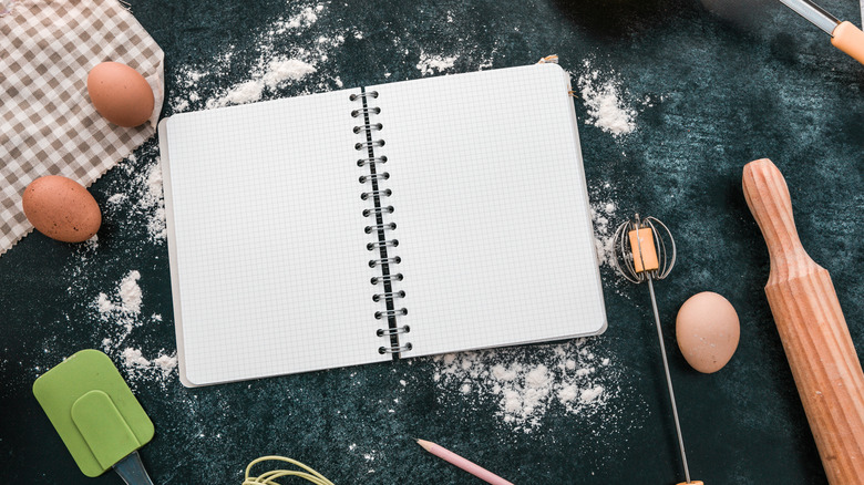 A blank notebook sits on a dark surface surrounded by cooking tools and ingredients