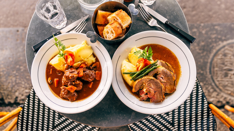 Two bowls of beef stew dishes sit side by side on a table with some crusted bread
