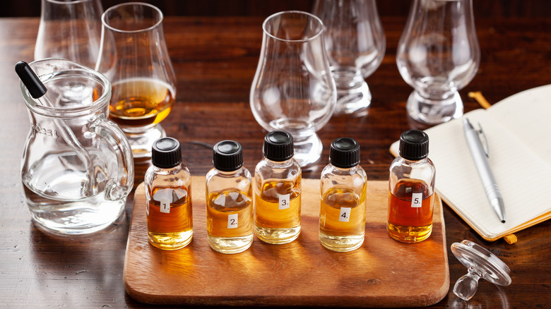 A whiskey tasting with five sample bottles, tasting glasses, a beaker of water with a dropper, and a notebook with a pen