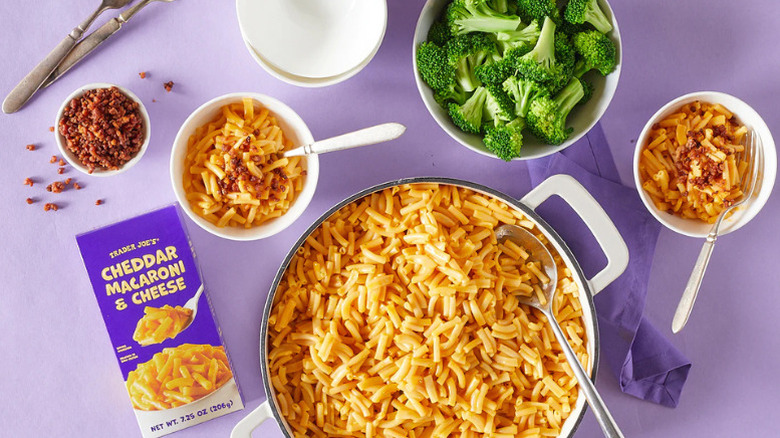 A pot of mac and cheese next to a bowl of bacon bits, two bowls of mac and cheese topped with bacon bits, a bowl of broccoli florets, and a box of Trader Joe's Cheddar Macaroni & Cheese