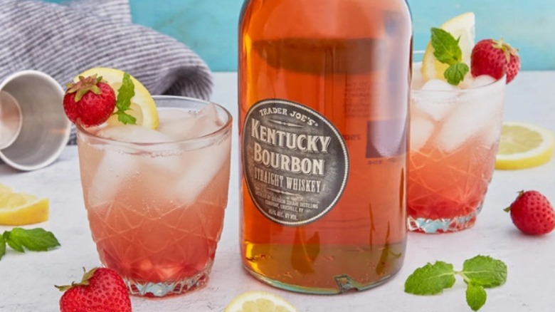 A bottle of Trader Joe's Kentucky bourbon sits in between two pink cocktails garnished with strawberries, lemons, and herbs