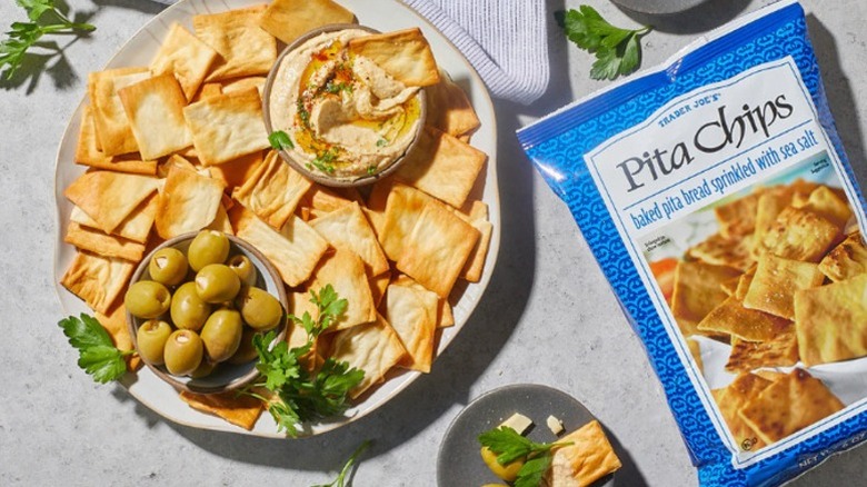 A plate of pita chips with small bowls of olives and hummus, with a parsley garnish, next to a bag of Trader Joe's Pita Chips