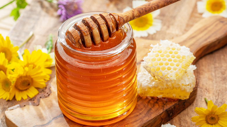 Honey in a glass jar with a honey dipper and honey comb on a wooden board