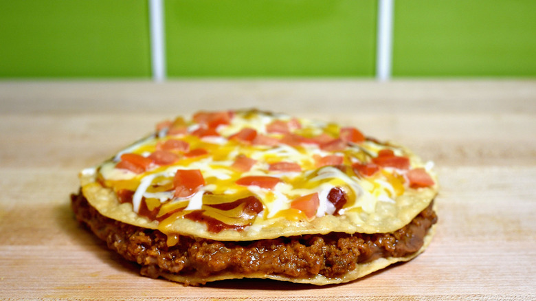 a Taco Bell Mexican Pizza featuring beef and beans between two tostada shells with melted red enchilada sauce, cheese, and tomatoes on top