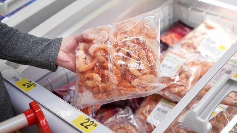 A person lifts a package of frozen shrimp out of a grocery store freezer.