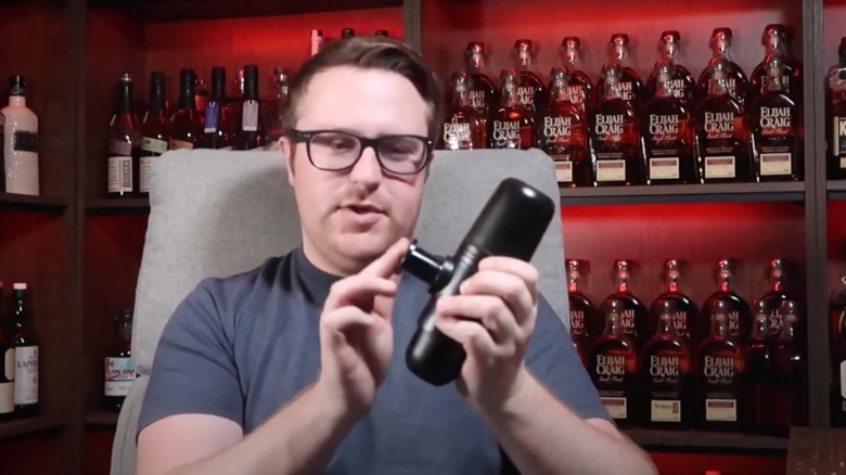 A man sits in a chair holding a whiskey pump, with multiple bottles of whiskey lining the walls behind him