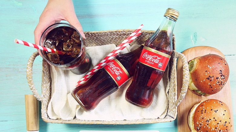 Bottles of Coca-Cola Ginger in a basket, with a glass and some picnic food.