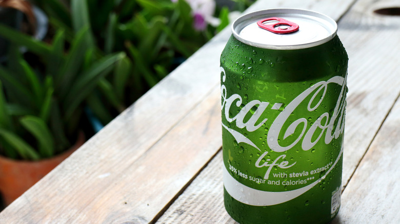 A green can of Coca-Cola Life on a wooden table with plants in the background.