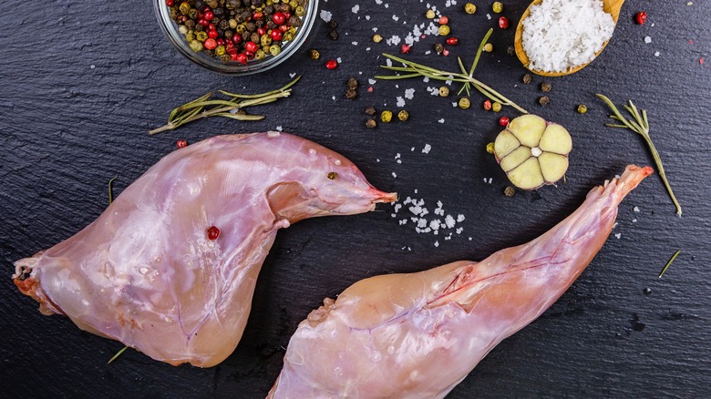 Close up of rabbit legs on black slate surface with salt, peppercorns, and raw garlic