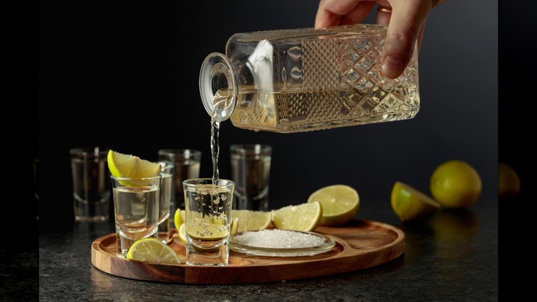 A person pouring tequila into a shot glass on a tray surrounded by limes and salt