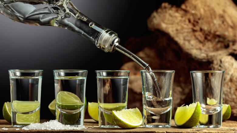 A spouted bottle pouring tequila into a lineup of shot glasses surrounded by salt and limes