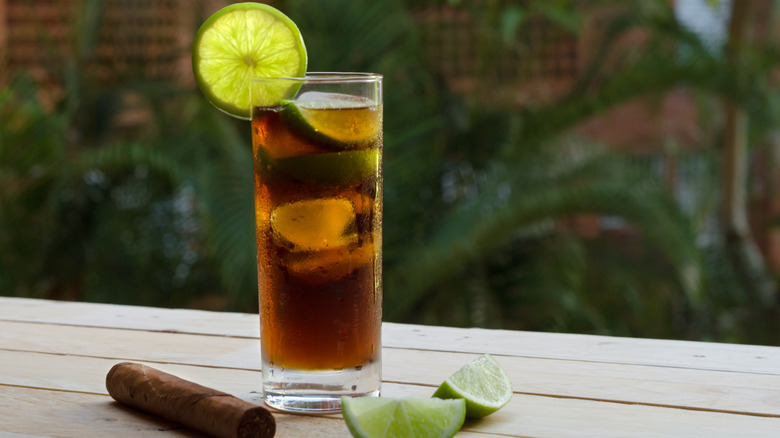 A Cuba Libre cocktail surrounded by sliced limes and a cigar