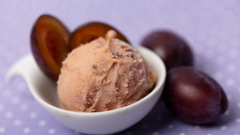 A scoop of plum ice cream in white bowl surrounded by plums